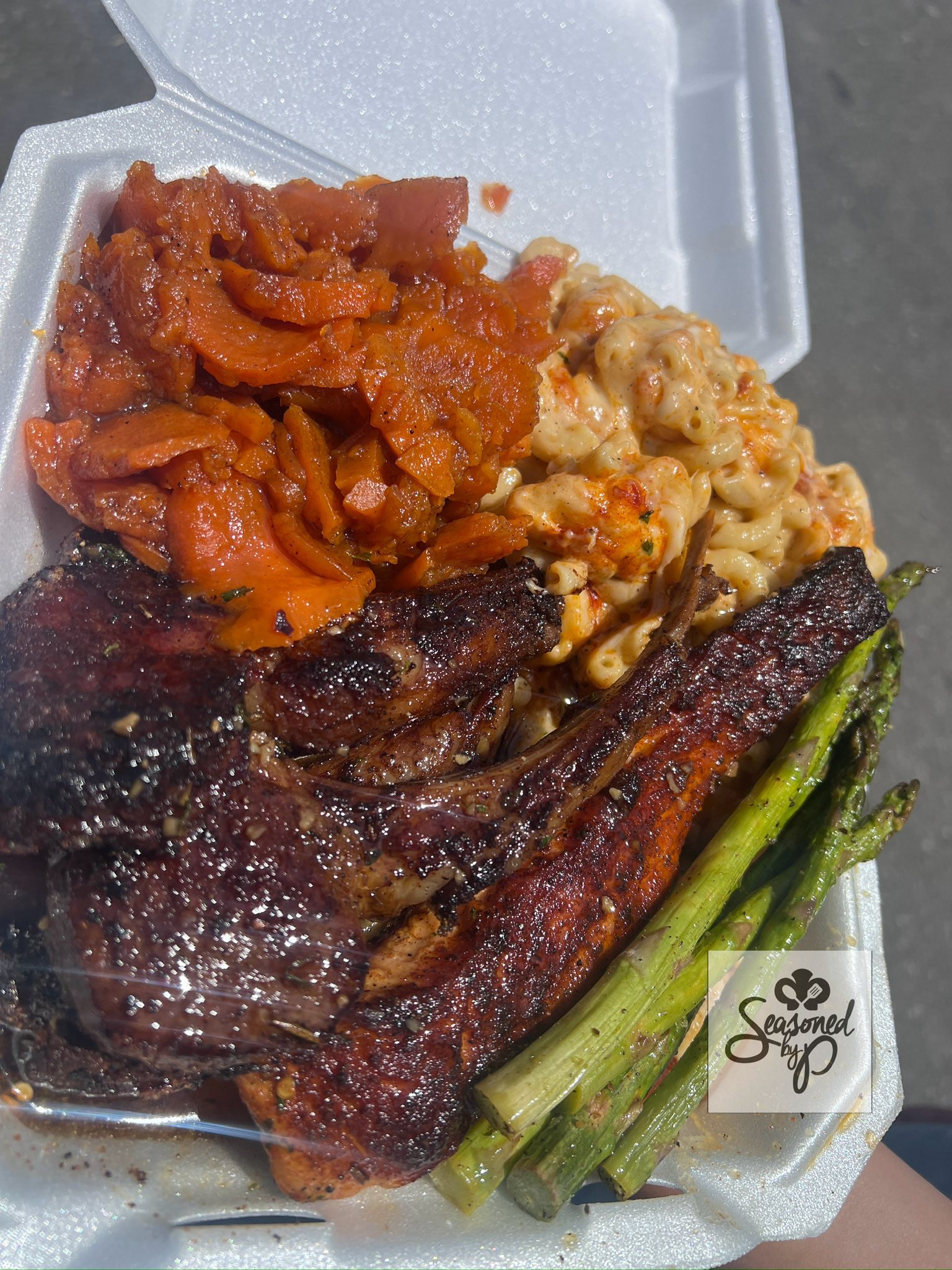 A takeout box of smothered chops and ribs beside candied yams, seafood mac and cheese and grilled asparagus