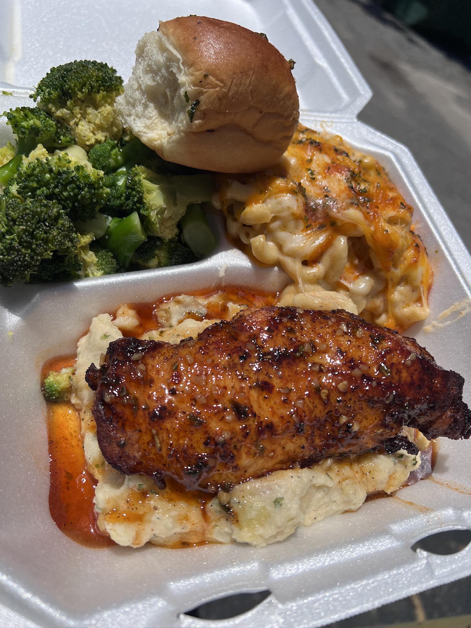Golden seasoned chicken breast resting on garlic mashed potatoes beside baked mac and cheese, broccoli and a buttered roll
