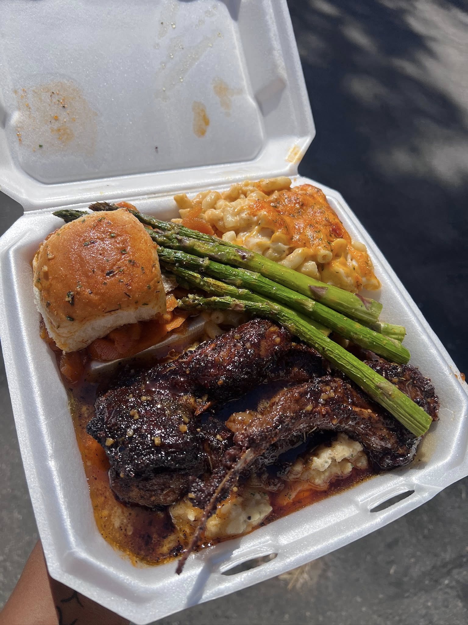 Smothered ribs and steak in a takeout box with roasted asparagus, baked mac and cheese and a garlic roll